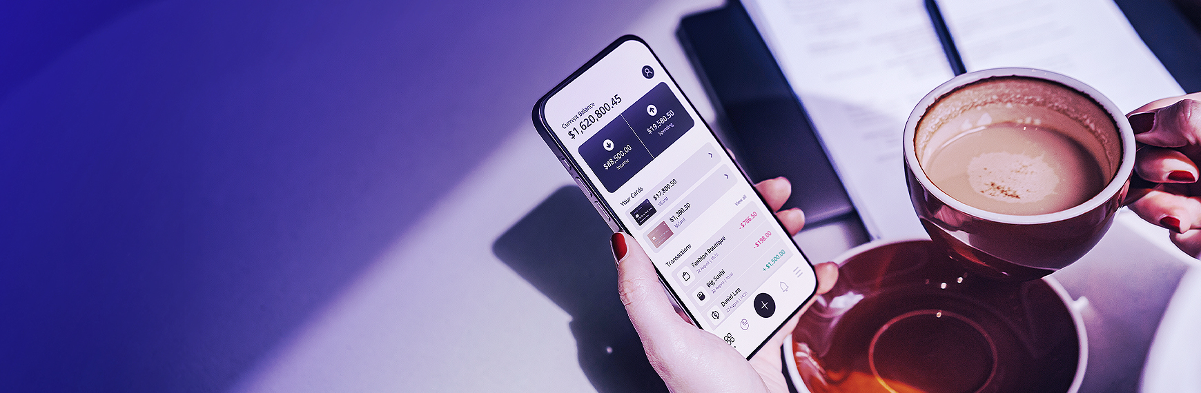 Symbolbild zu CCDII: Hand hält Handy mit Bank-App in der Hand; daneben Kaffeetasse